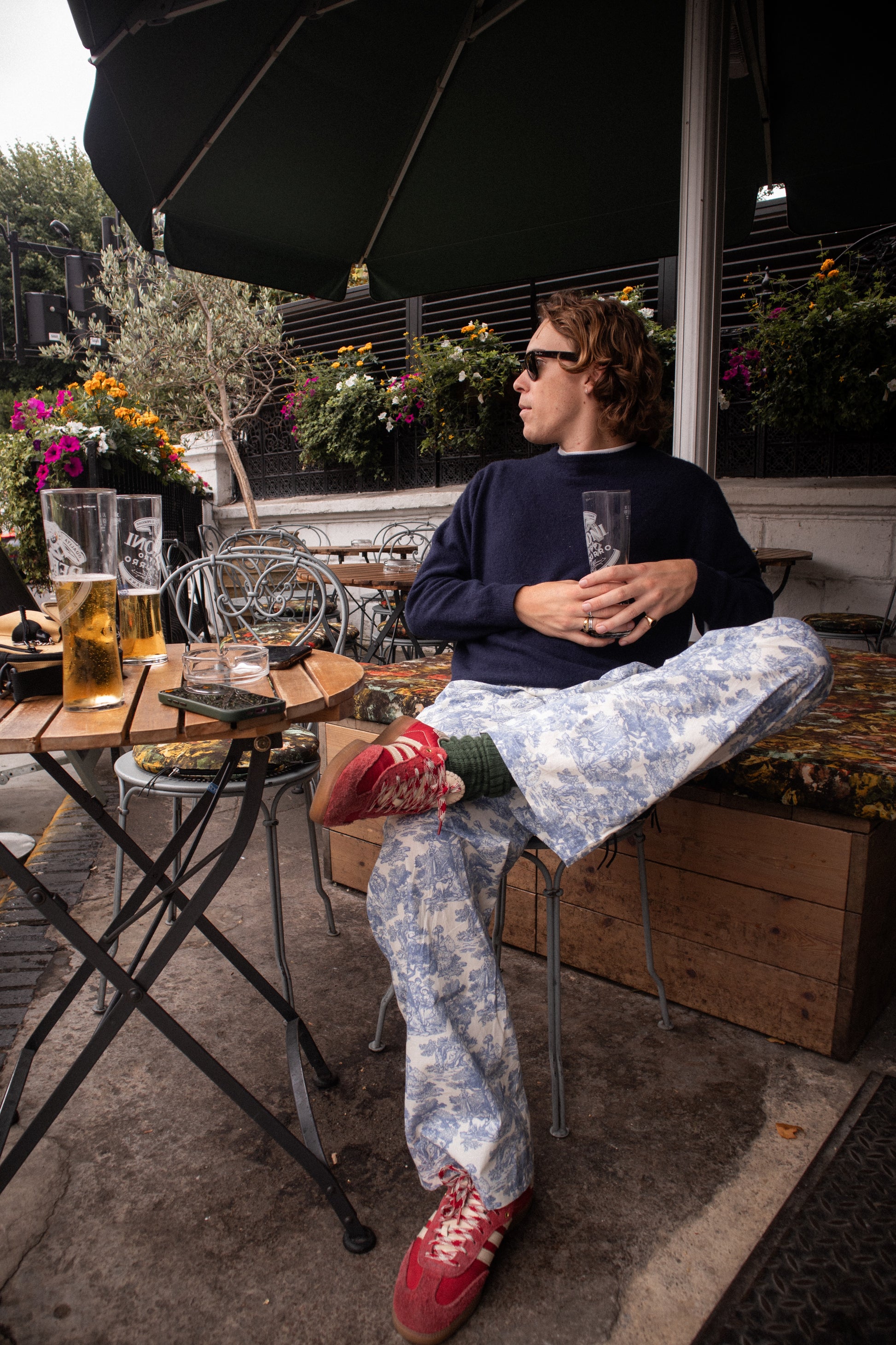 Person sitting at an outdoor table with drinks, surrounded by plants and flowers.